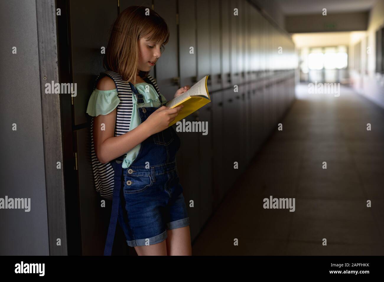 Scolaretta appoggiata contro un muro in un corridoio esterno che legge un libro alla scuola elementare Foto Stock