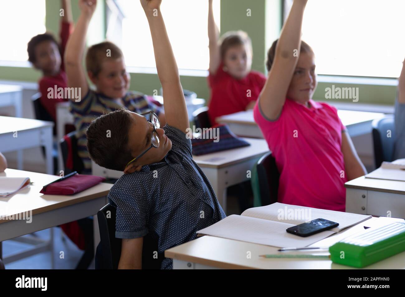 Gruppo di bambini che siedono alle scrivanie e alzano le mani per rispondere Foto Stock