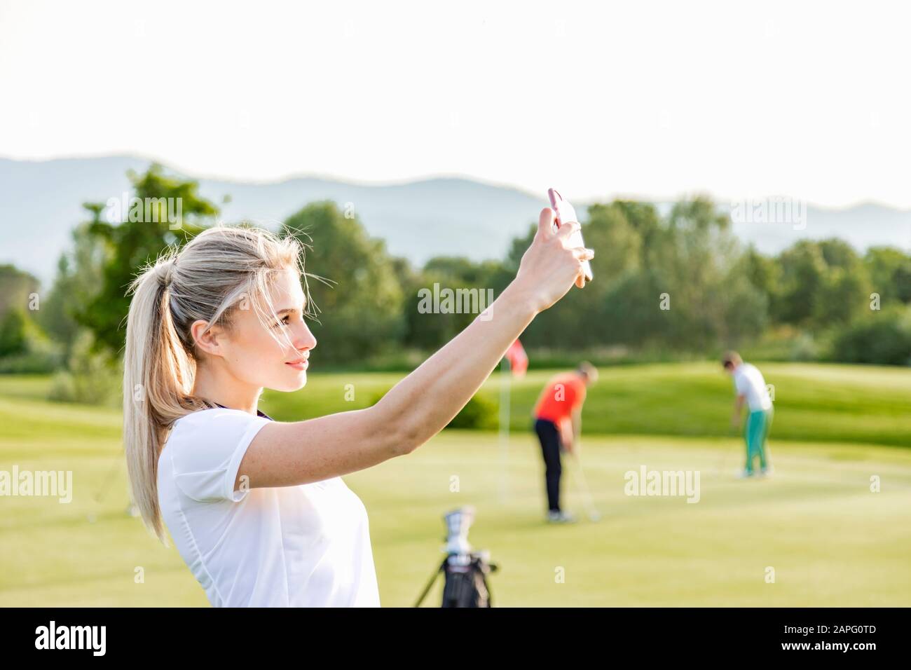 Donna che prende selfie sul campo da golf, amici che giocano a golf in background Foto Stock