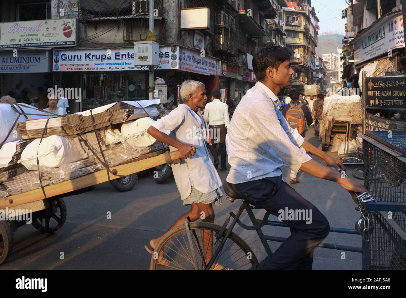 Nella trafficata Kalbadevi Road, Bhuleshwar, Mumbai, India, un portiere lotta per tirare il suo pesante carrello attraverso il traffico caotico, sorvolato da un ciclista Foto Stock