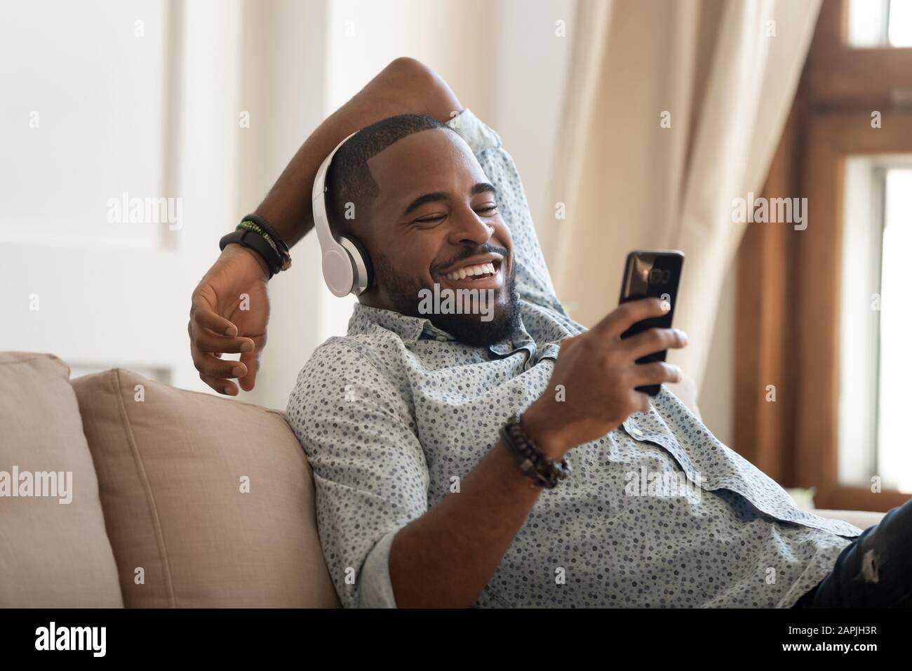 Ragazzo africano in cuffie appoggiato sul divano utilizzando lo smartphone ridere Foto Stock