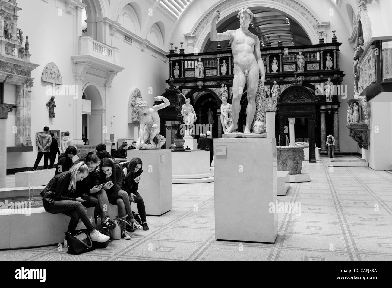 Fotografia in bianco e nero di Londra: Un gruppo di ragazze adolescenti studia i loro smartphone nella galleria di sculture classiche del V&A Museum, Londra, Regno Unito Foto Stock