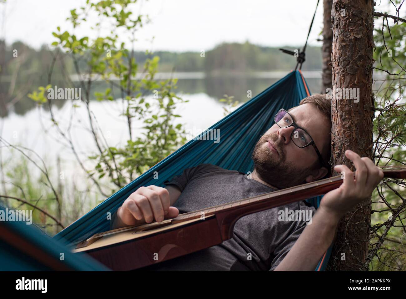 L'uomo triste si trova in un'amaca con una chitarra acustica in mano, suona una melodia e guarda sognatamente al cielo, sulla riva di un lago. Foto Stock