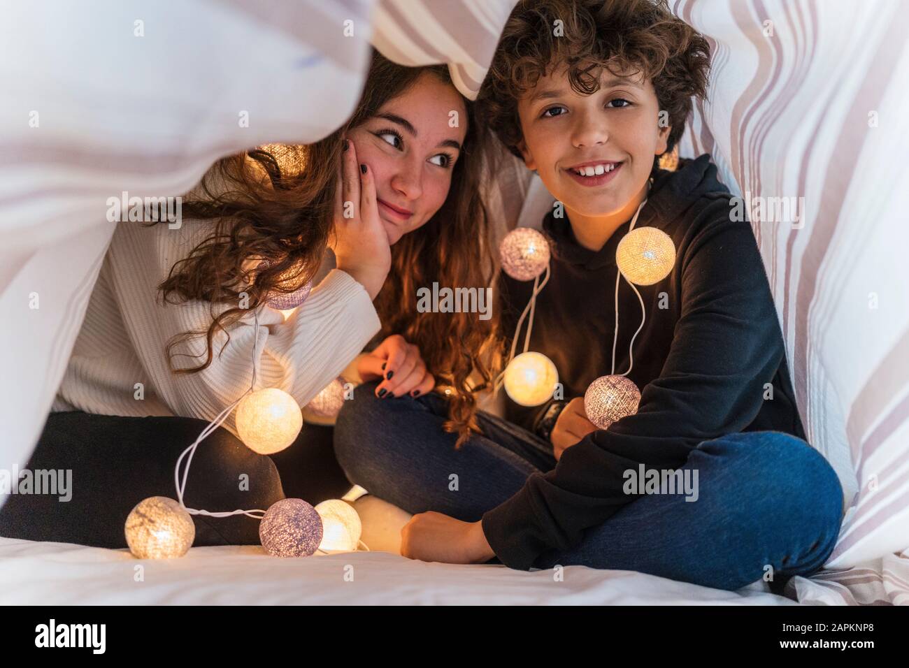 Ritratto di fratello felice e sorella con catena di luci sotto la copertura del letto Foto Stock