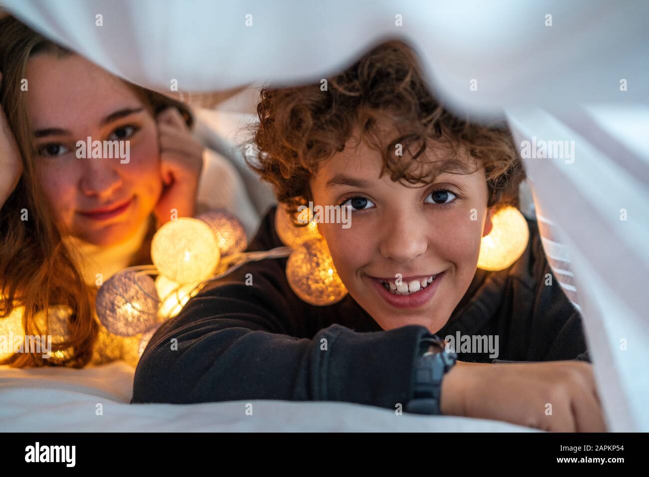 Ritratto di fratello felice e sorella con catena di luci sotto la copertura del letto Foto Stock