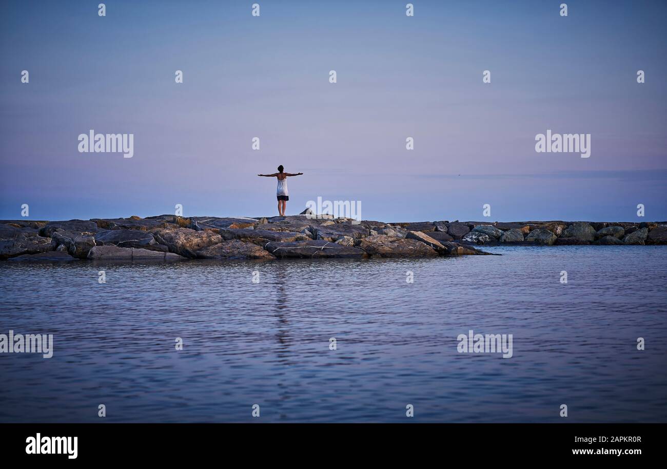 Vista posteriore della donna matura in piedi su rocce di fronte al mare al crepuscolo, Italia Foto Stock