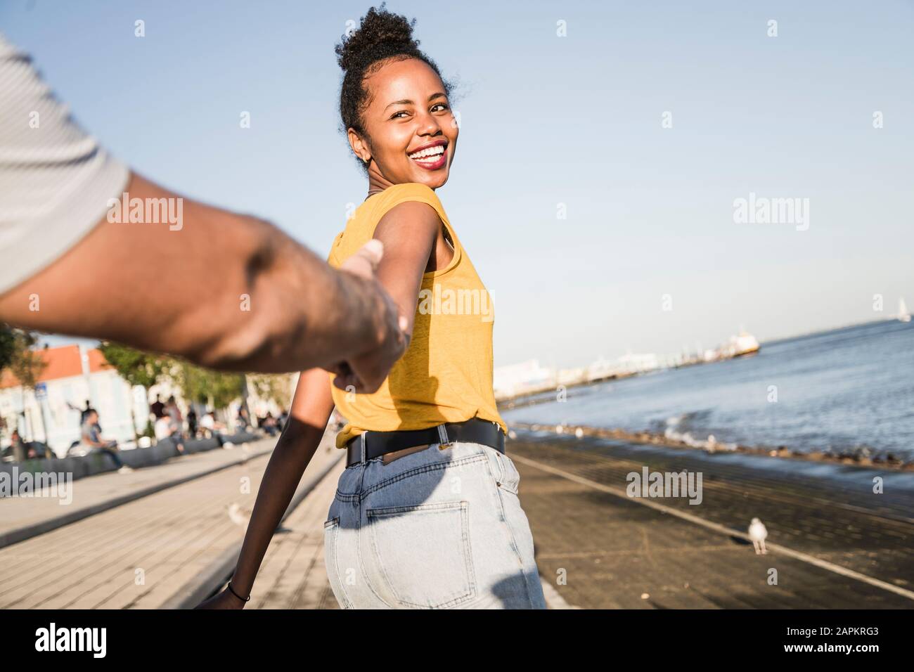 Felice giovane donna che tiene la mano del suo ragazzo sul molo sul lungomare, Lisbona, Portogallo Foto Stock