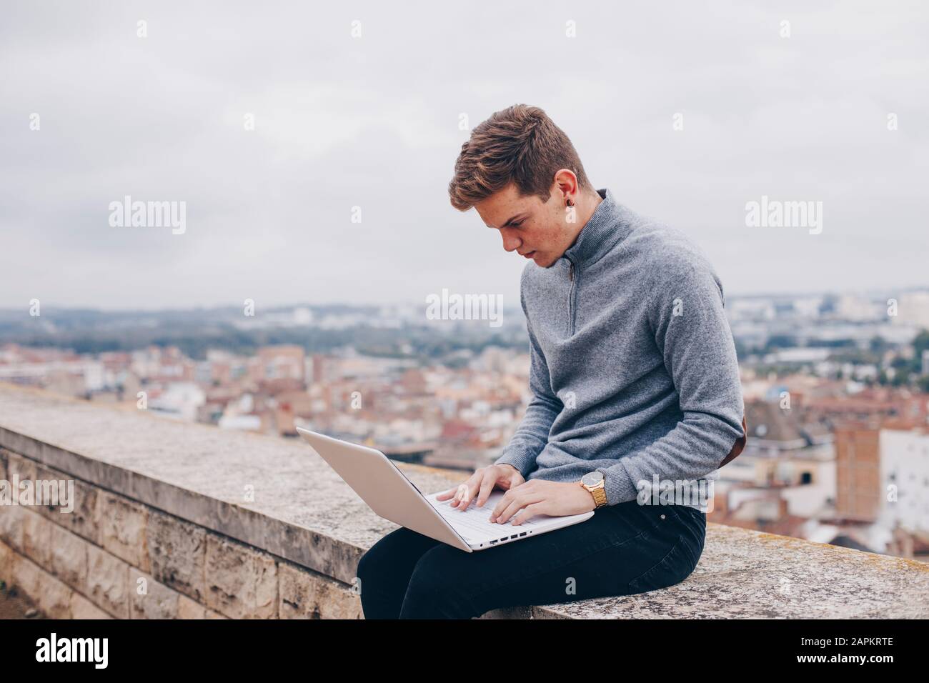 Giovane biondo seduto su un muro e digitando sul suo portatile a Lleida Foto Stock