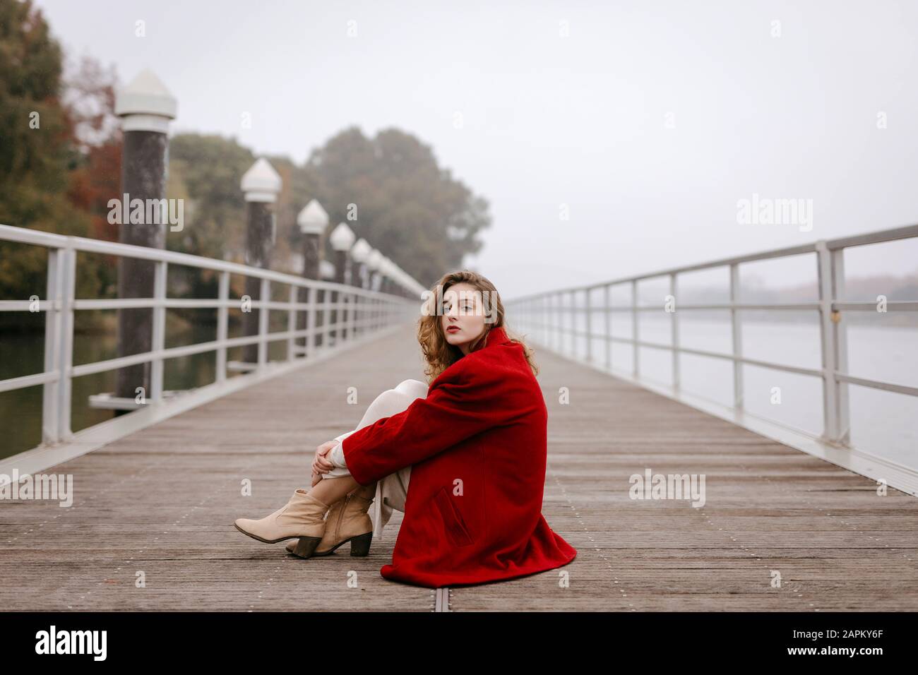 Ritratto di giovane donna che indossa cappotto rosso, appoggiato sulla ringhiera in giornata piovosa Foto Stock