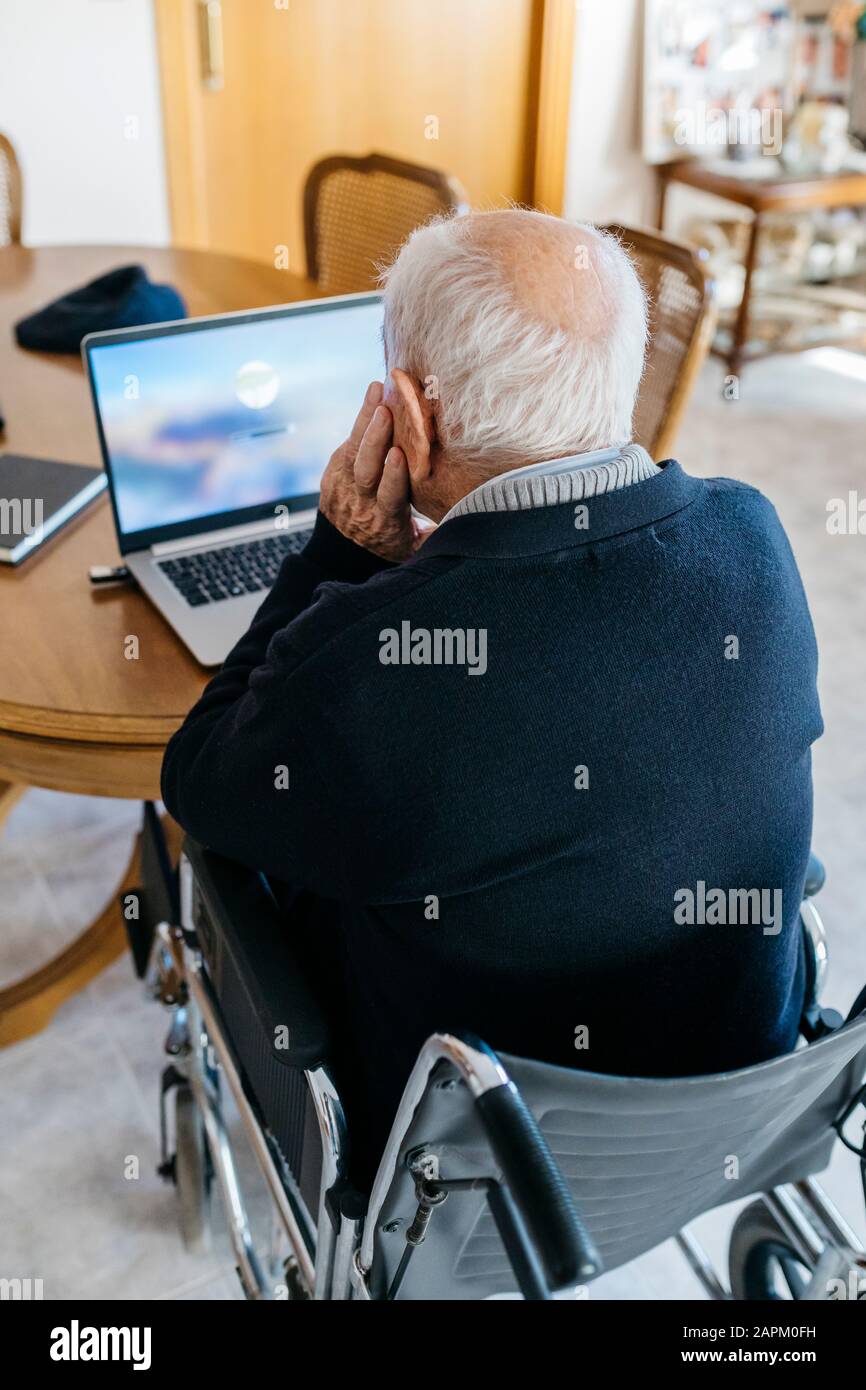Vista posteriore dell'uomo anziano in sedia a rotelle utilizzando il laptop a casa Foto Stock