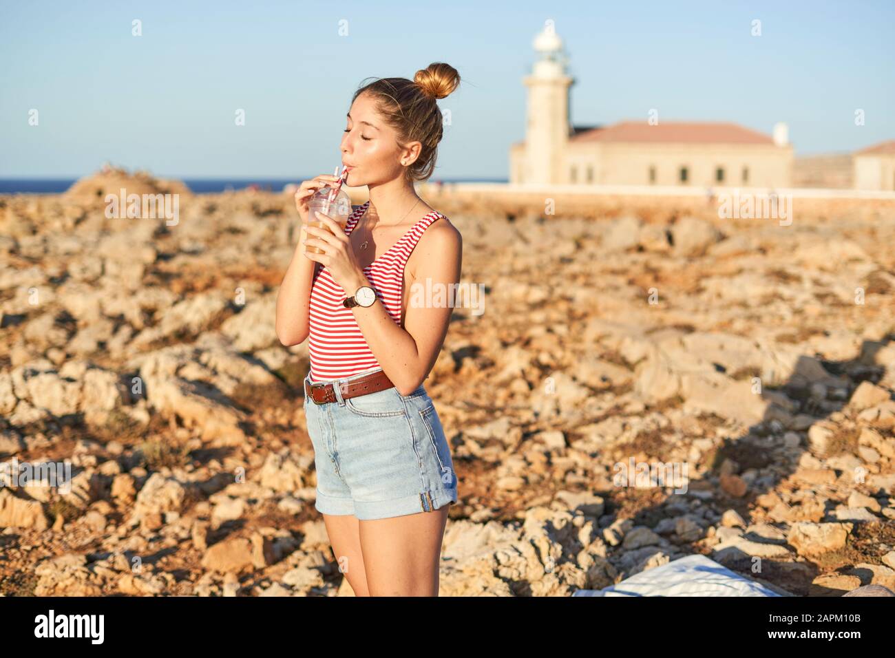 Giovane donna che beve succo sulla spiaggia rocciosa, Minorca, Spagna Foto Stock
