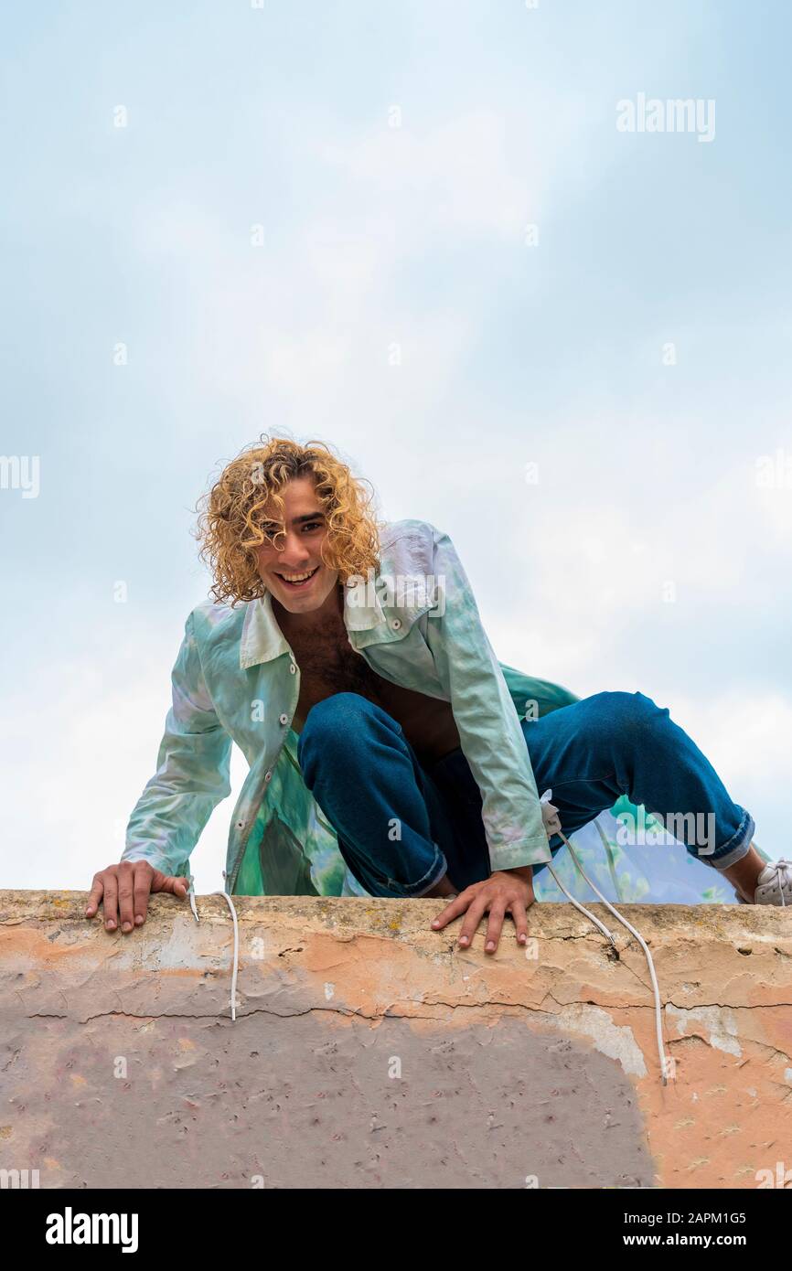 Giovane biondo sorridente su un muro, guardando la macchina fotografica Foto Stock