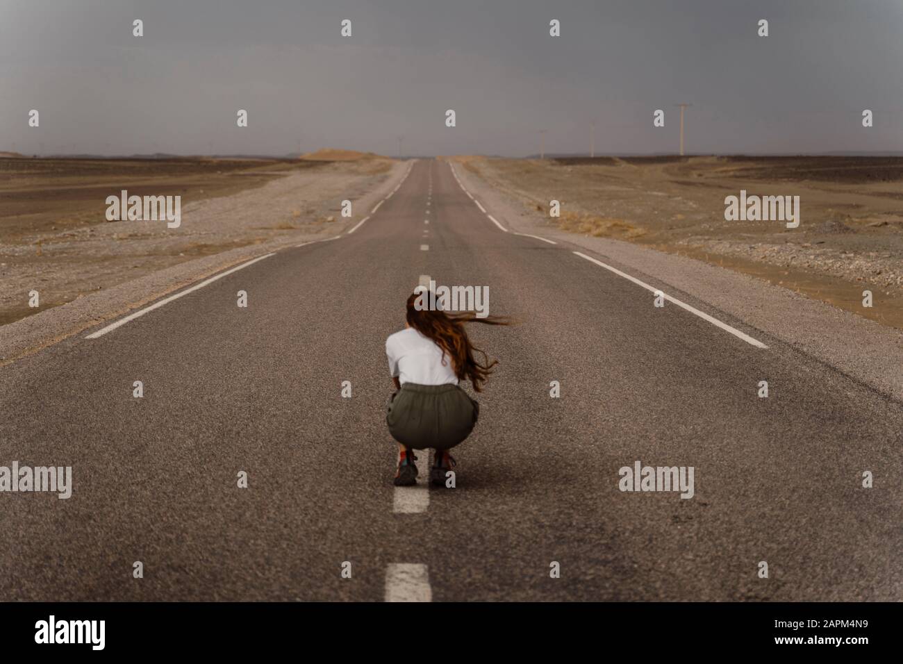 Vista posteriore della donna accovacciata sulla striscia mediana della strada di campagna vuota, Fez, Marocco Foto Stock