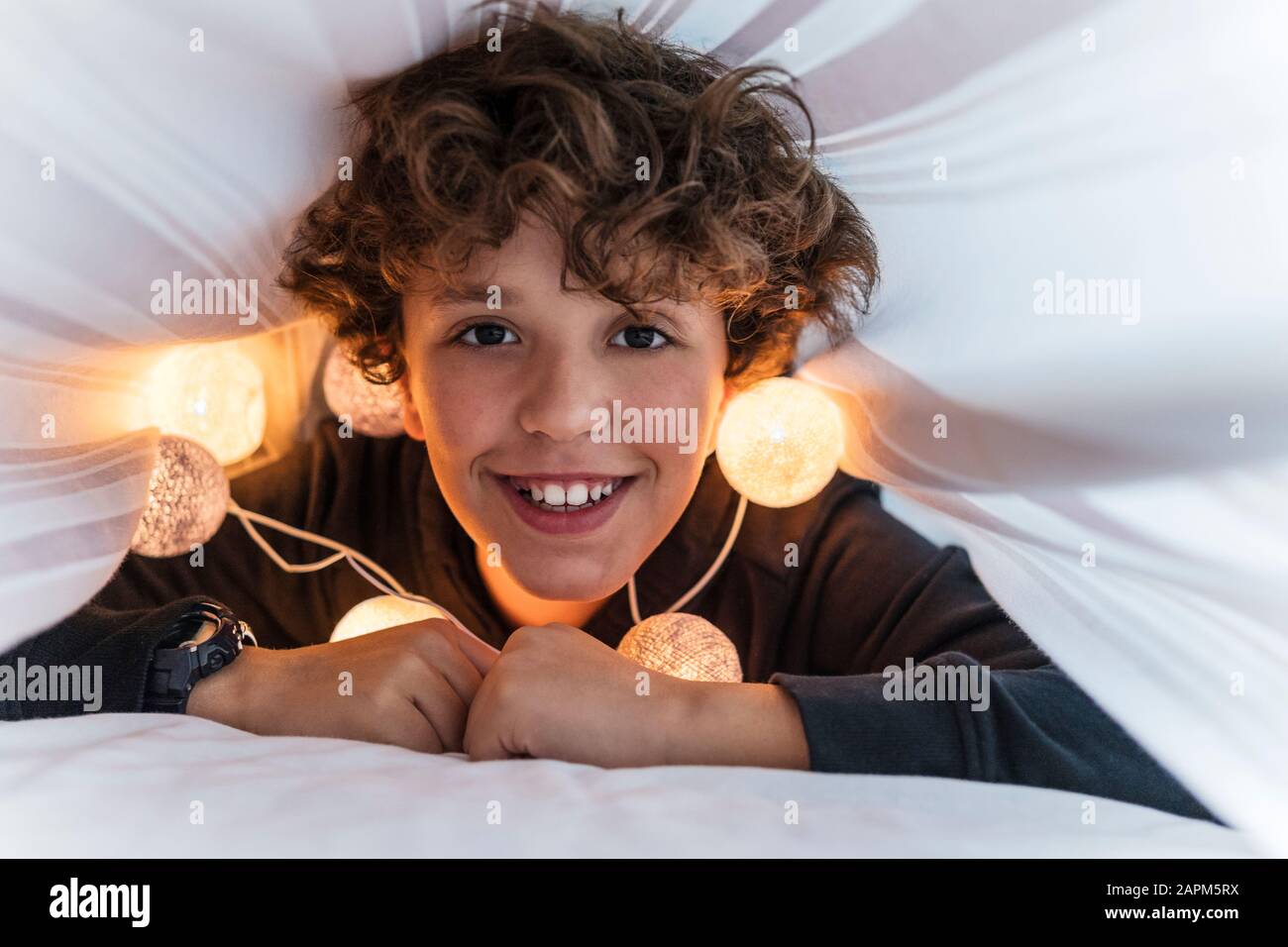 Ritratto di ragazzo felice con una catena di luci sotto il copriletto Foto Stock