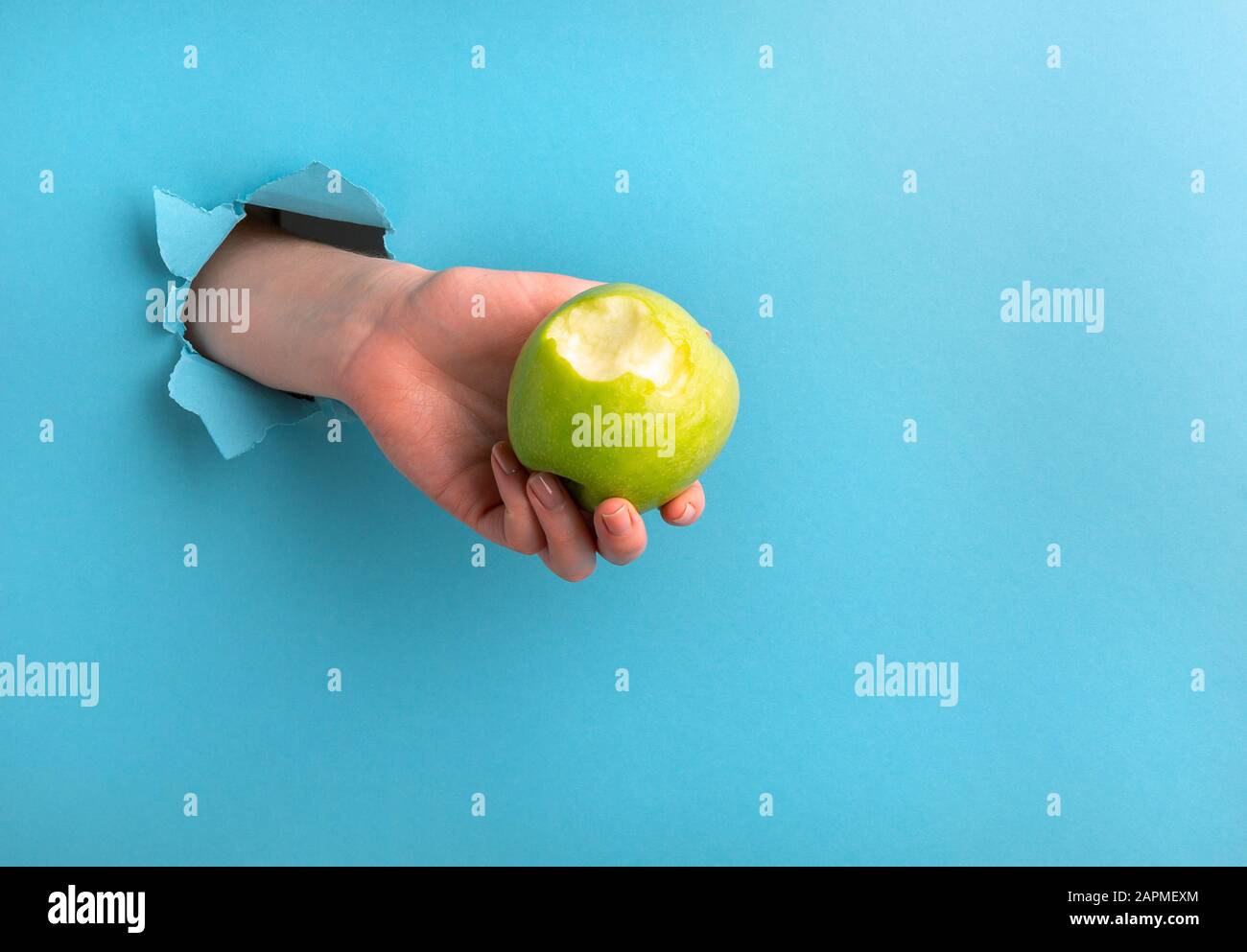 La mano di una donna tiene una mela bitten attraverso un buco nella carta su uno sfondo blu. Spazio di copia Foto Stock