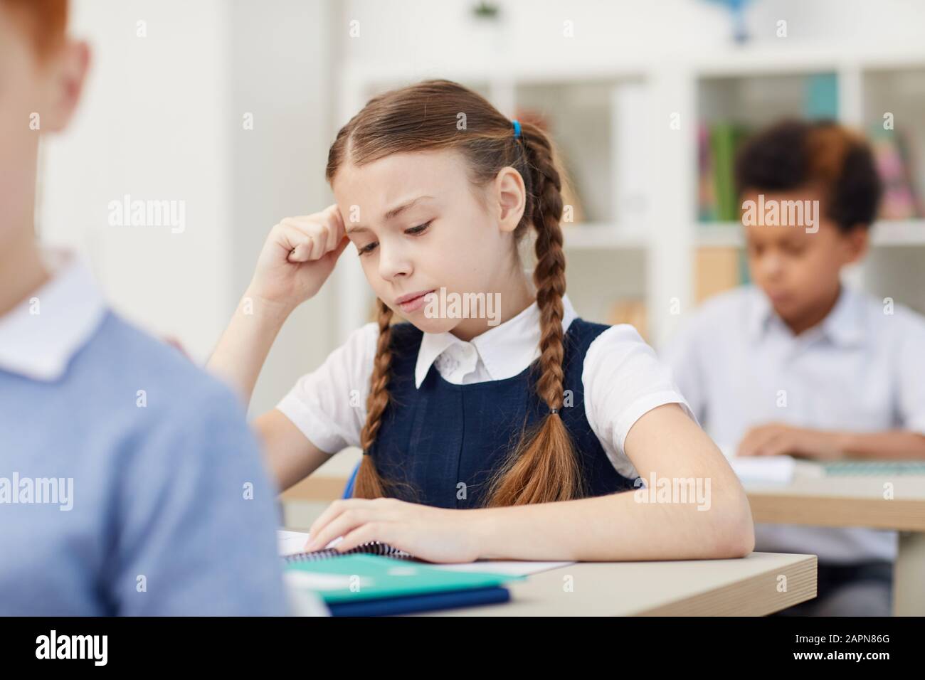 Una studentessa seria seduta alla scrivania e studiando durante una lezione con gli altri bambini della scuola in classe Foto Stock