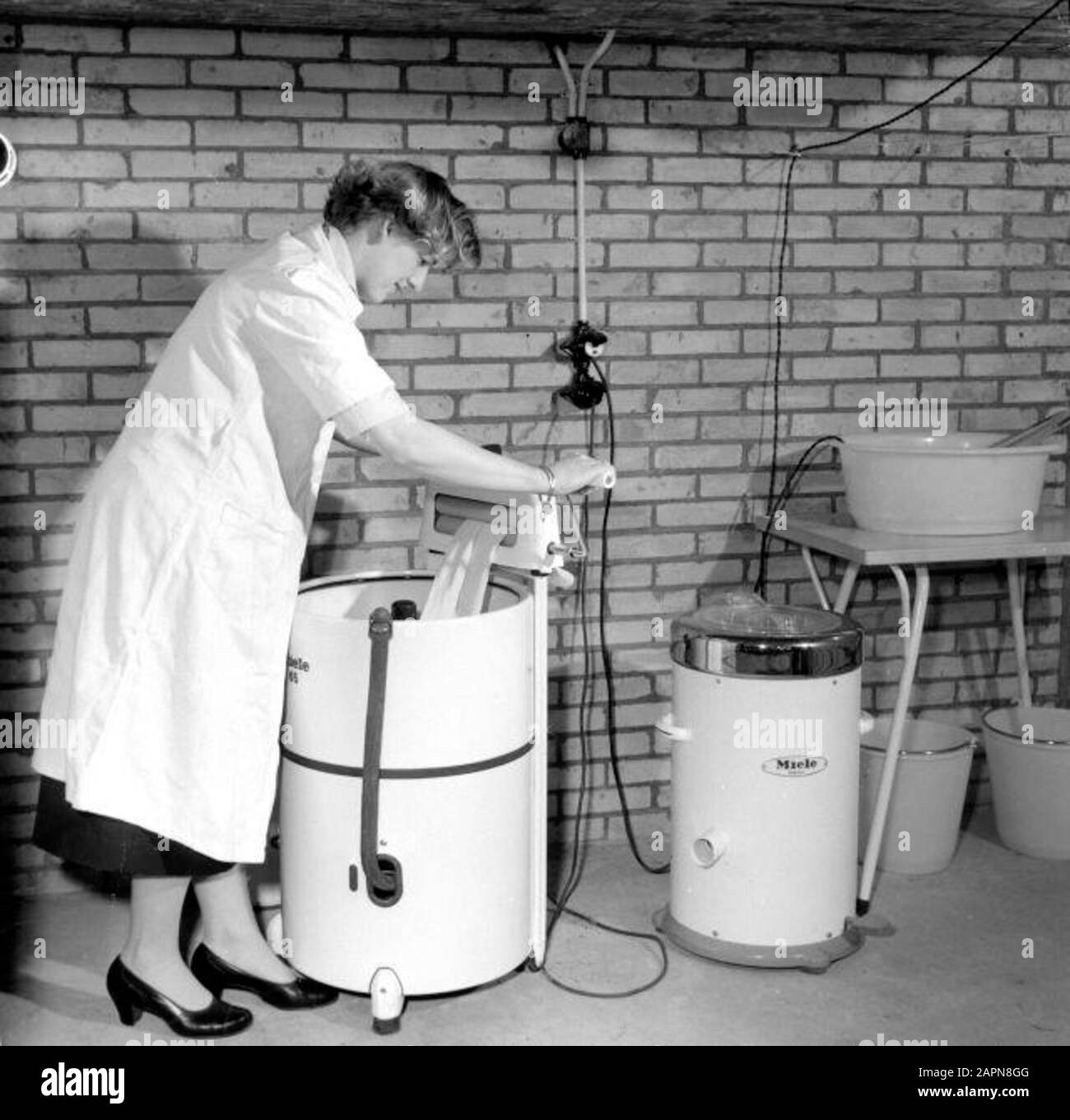 Lavanderia giorno alla famiglia Bex a Maarssen. Signora Bex in un grembiule nel garage o nella sala di servizio. Lei tira la cera attraverso lo strizzatore sulla sua Miele lenta lavatrice, accanto ad essa una centrifuga Miele. Febbraio 1957. Foto Stock