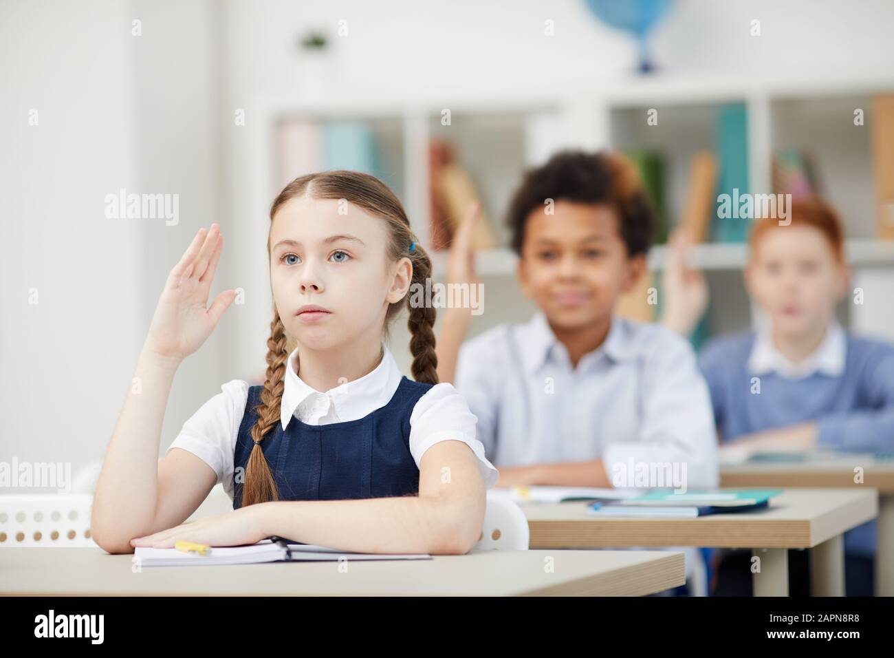 Una studentessa seria seduta alla scrivania e alzando la mano con altri compagni di classe in background durante una lezione a scuola Foto Stock