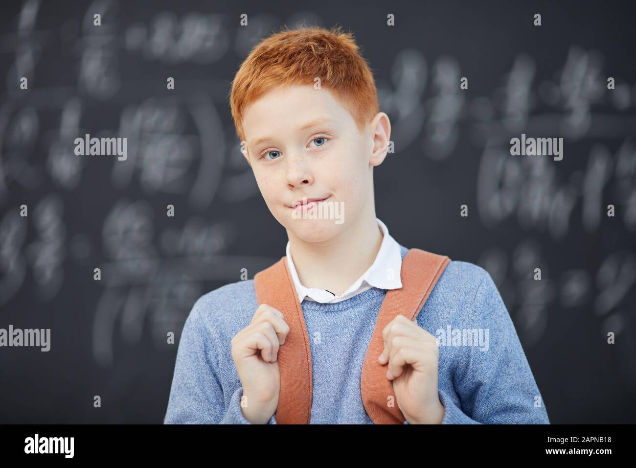 Ritratto di scolaro rosso con zaino che guarda la macchina fotografica con lavagna sullo sfondo è a lezione di matematica Foto Stock