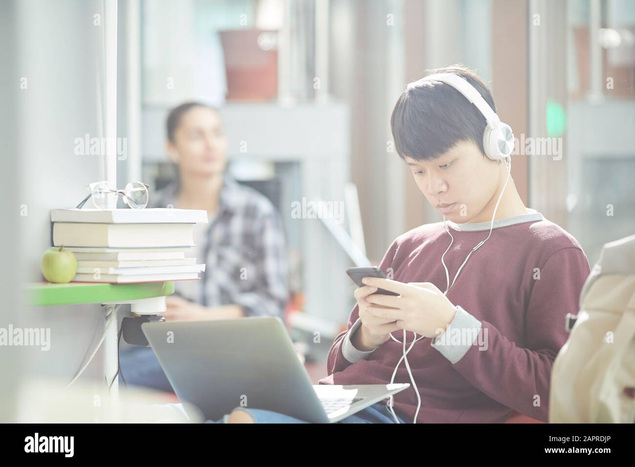 Studente di scuola asiatica in cuffie seduti sul divano con il computer portatile in ginocchio e utilizzando il telefono cellulare Foto Stock