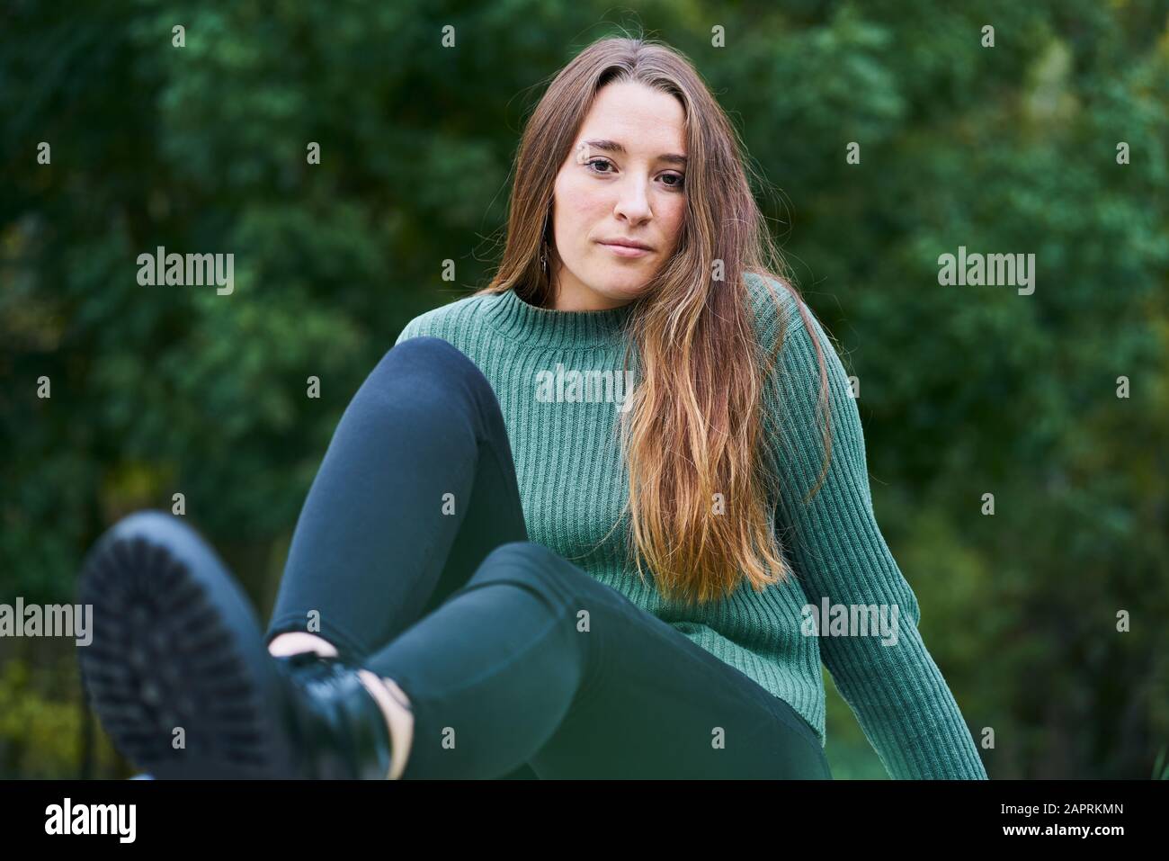 Ritratto di una giovane donna caucasica con capelli castani in un parco con il verde degli alberi sullo sfondo Vestito da maglione verde, pantaloni scuri e Foto Stock