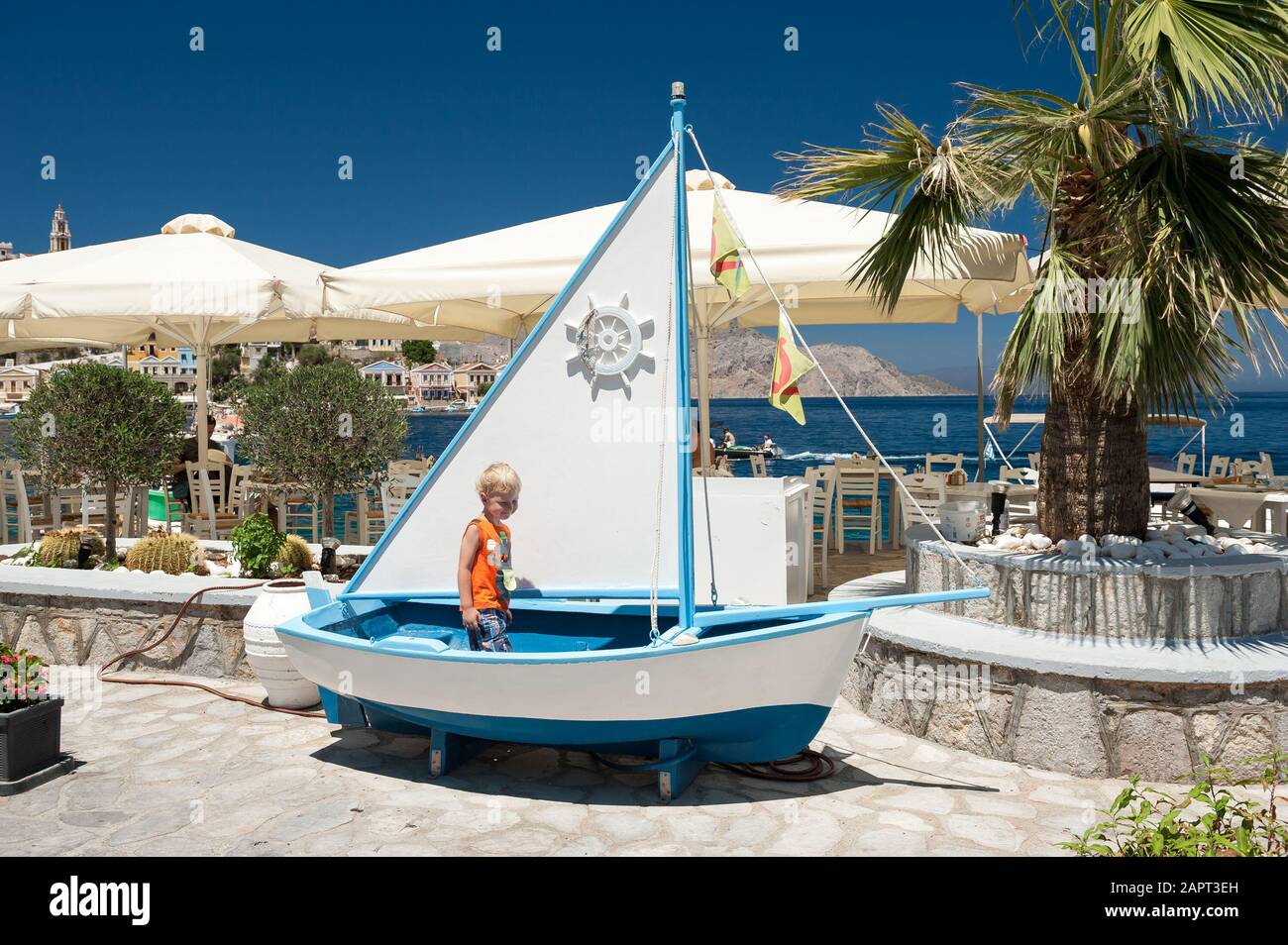 Tre anni di old boy in piedi in una barca di fronte ad un ristorante in Symi, Grecia, Europa Foto Stock