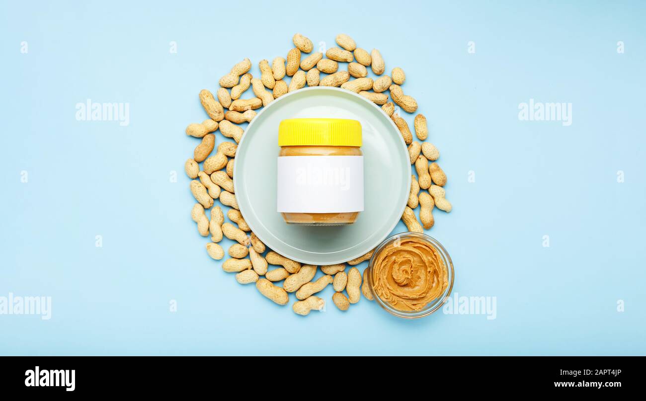Pasta di arachidi cremosa in vaso di vetro con tappo giallo con mock-up e burro di arachidi in una lastra di vetro. Arachidi nella buccia sparse su sfondo blu Foto Stock