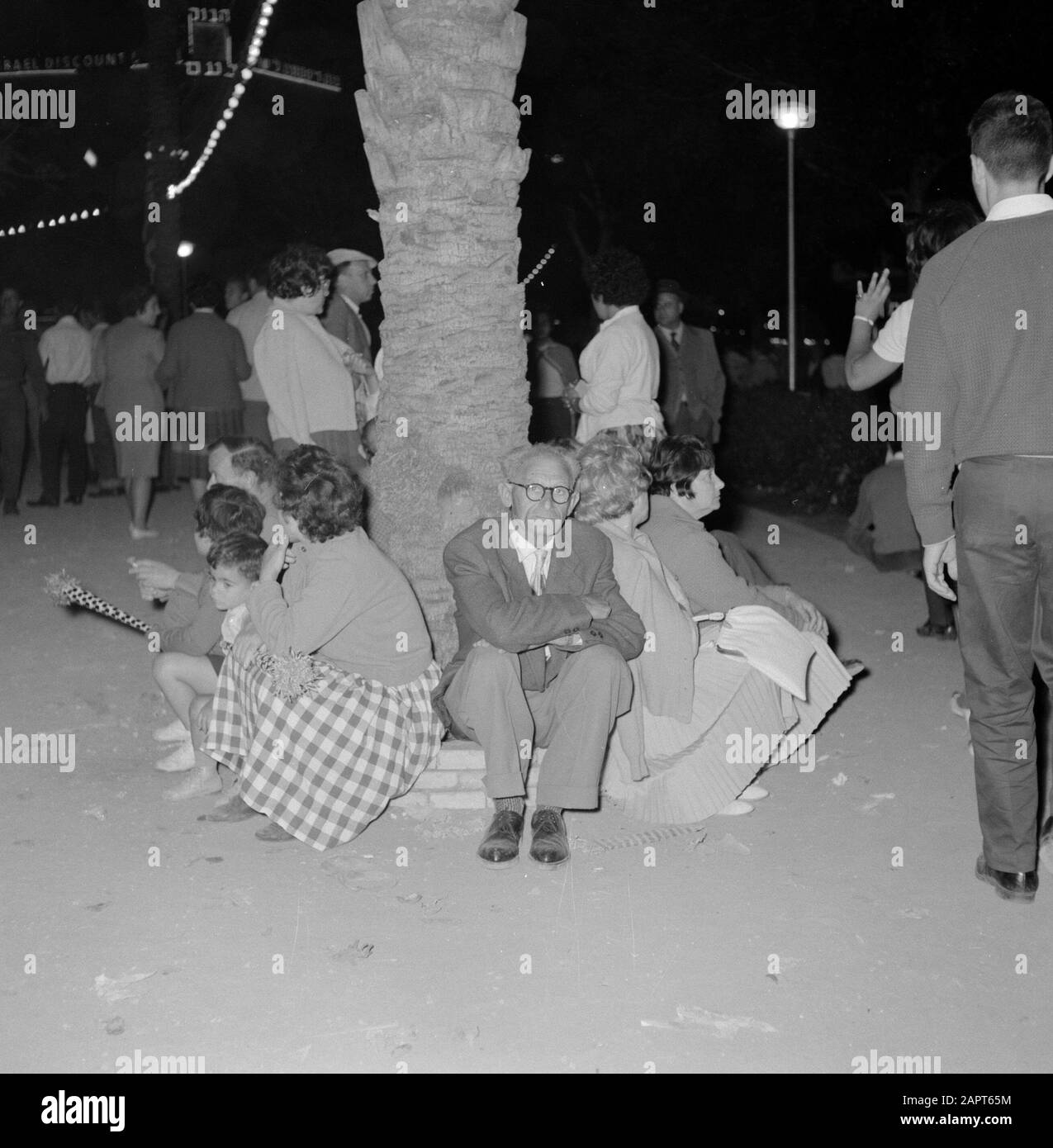 Israele 1964-1965: Tel Aviv, celebrazione dei Festaioli del giorno dell'indipendenza, siedono sul bordo intorno ad un albero e riposano mentre altri continuano a celebrare l'annotazione: I residenti celebrano il giorno dell'indipendenza dello Stato di Israele, ballano sulla strada, vendono e comprano i beni di festa e mangiano antipasti festosi Data: 1964 luogo: Israele, Tel Aviv Parole Chiave: Alberi, articoli di festa, immagini notturne, vacanze nazionali, immagini di strada Foto Stock