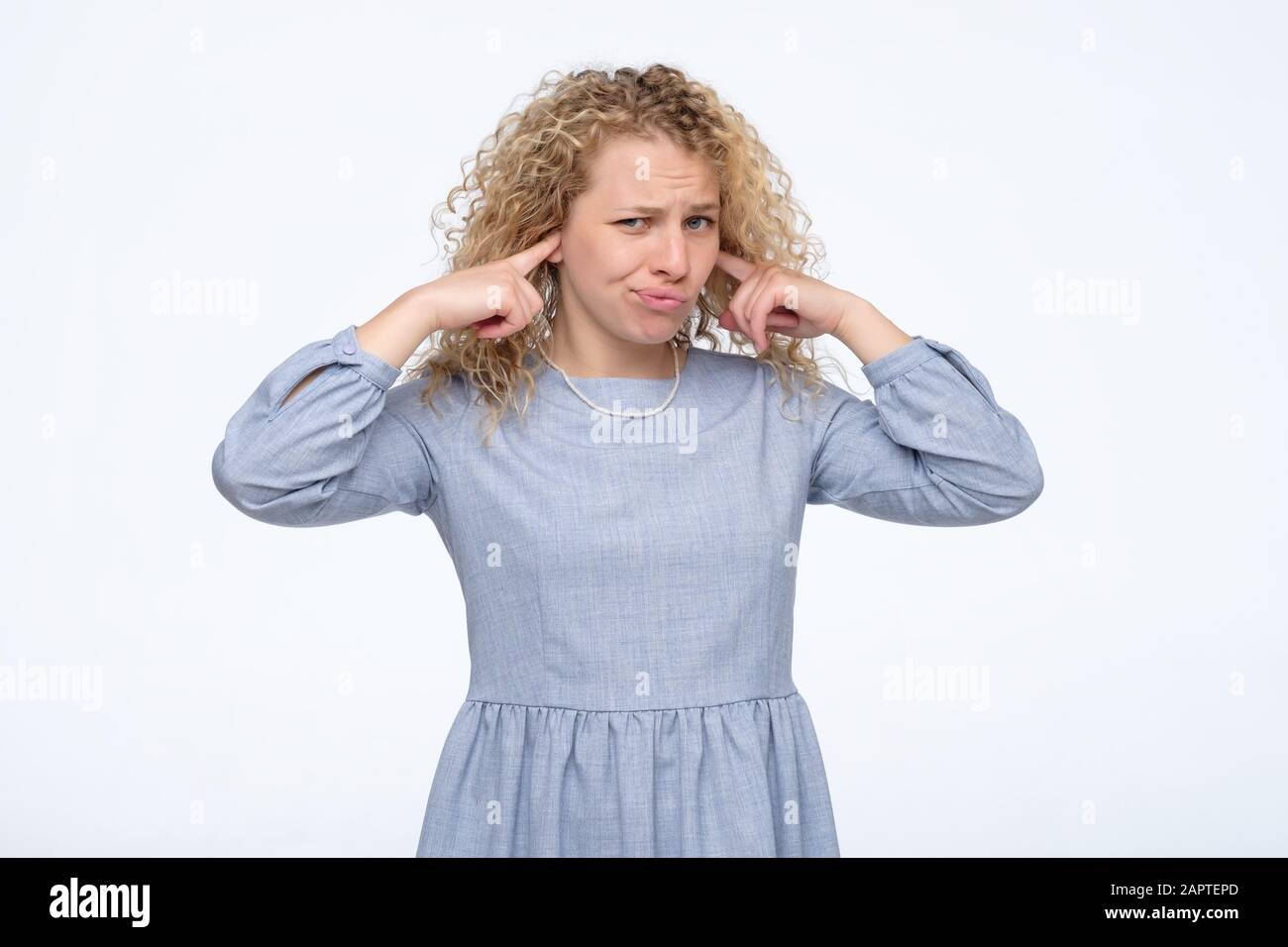 Giovane donna bionda curly in abito blu che copre le orecchie. Studio. Foto Stock