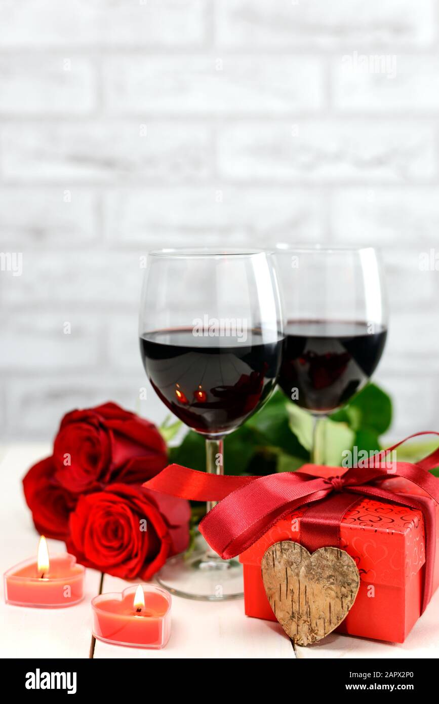 Concetto di San Valentino. Due bicchieri di vino, rose rosse, scatola regalo, cuore di legno e candele accese su un tavolo di legno bianco. Messa a fuoco selettiva. Foto Stock