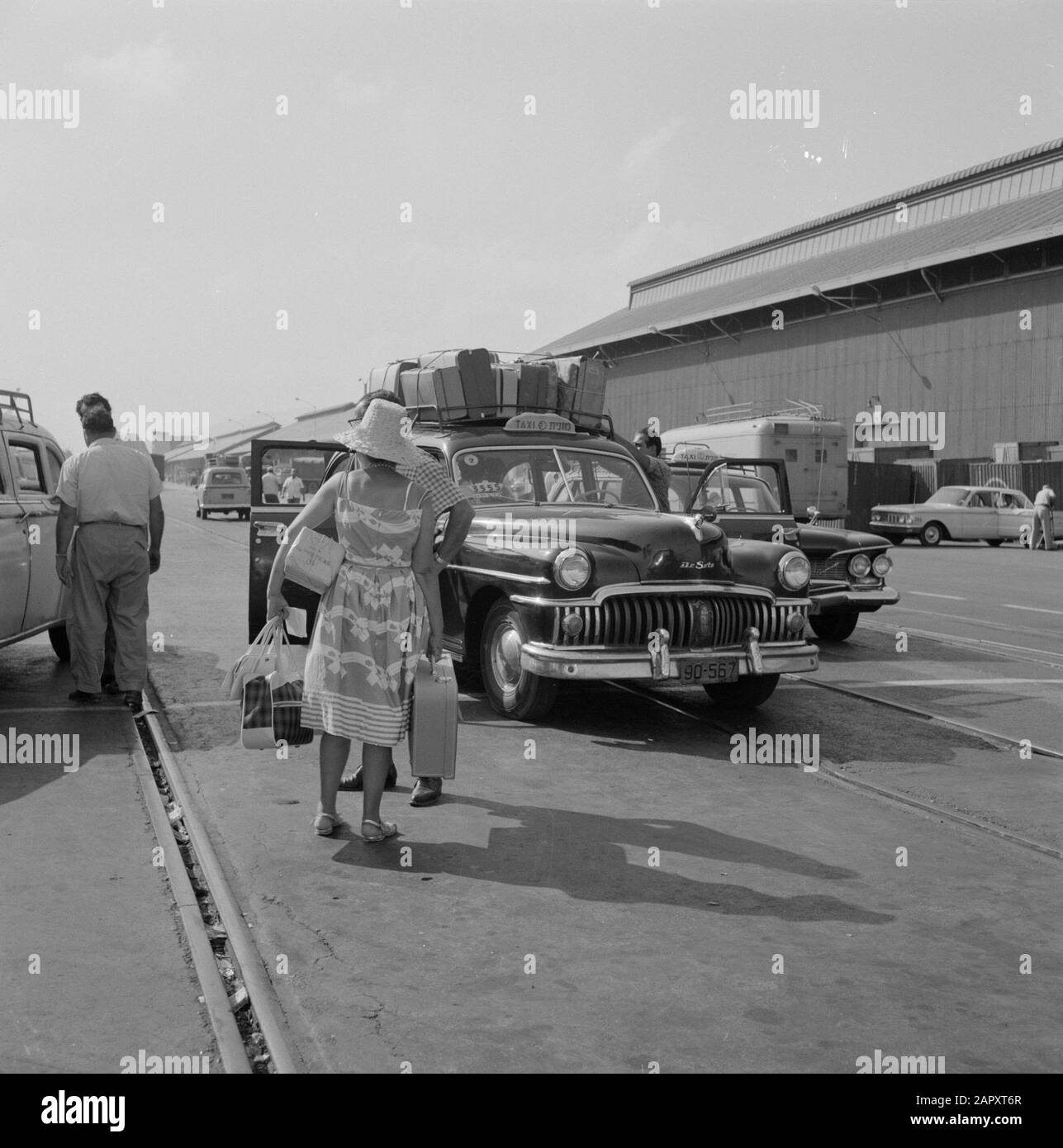 Israele 1964-1965: Haifa, arrivo di navi passeggeri Valige Legate sul tetto di un taxi e una donna vestito d'estate con le mani piene di bagagli nel parcheggio Annotazioni: Haifa è una città portuale e una località balneare nel nord di Israele, nella parte più settentrionale delle montagne del Carmelo e sul Mar Mediterraneo. Nel 1945, ZIM Lines (compagnia di navigazione) iniziò come mittente di immigrati ebrei nel nuovo stato di Israele ed è diventata una delle più grandi compagnie di navigazione del mondo con filiali in tutto il mondo Data: 1964 luogo: Haifa, Israele Parole Chiave: Arrivo e partenza, bagagli, porti, tra Foto Stock