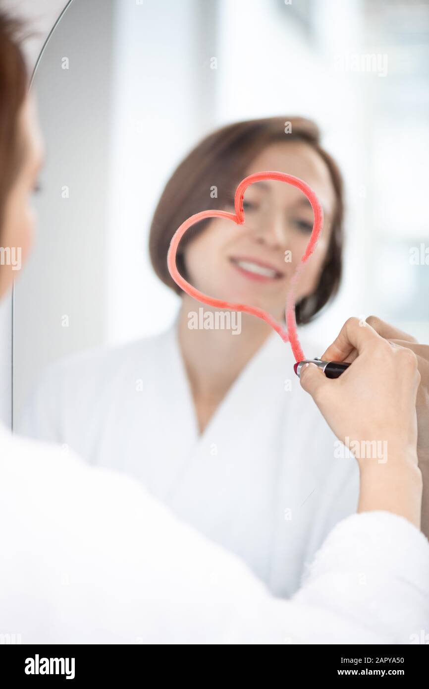 Riflesso di giovane donna flirty che disegna il cuore con rossetto cremisi sullo specchio Foto Stock