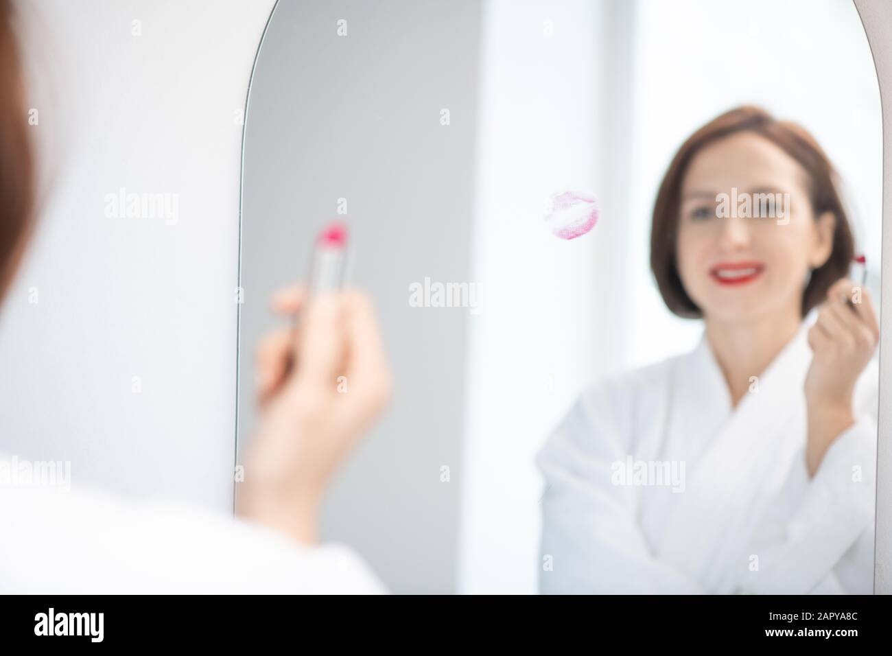Riflessione in specchio di giovane donna in accappatoio bianco che tiene rossetto cremisi Foto Stock