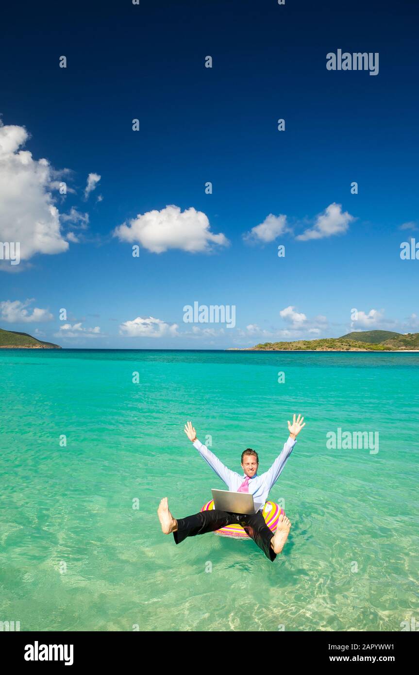 Uomo d'affari che lancia le braccia e le gambe in festa mentre galleggia in una baia tropicale con il suo portatile Foto Stock