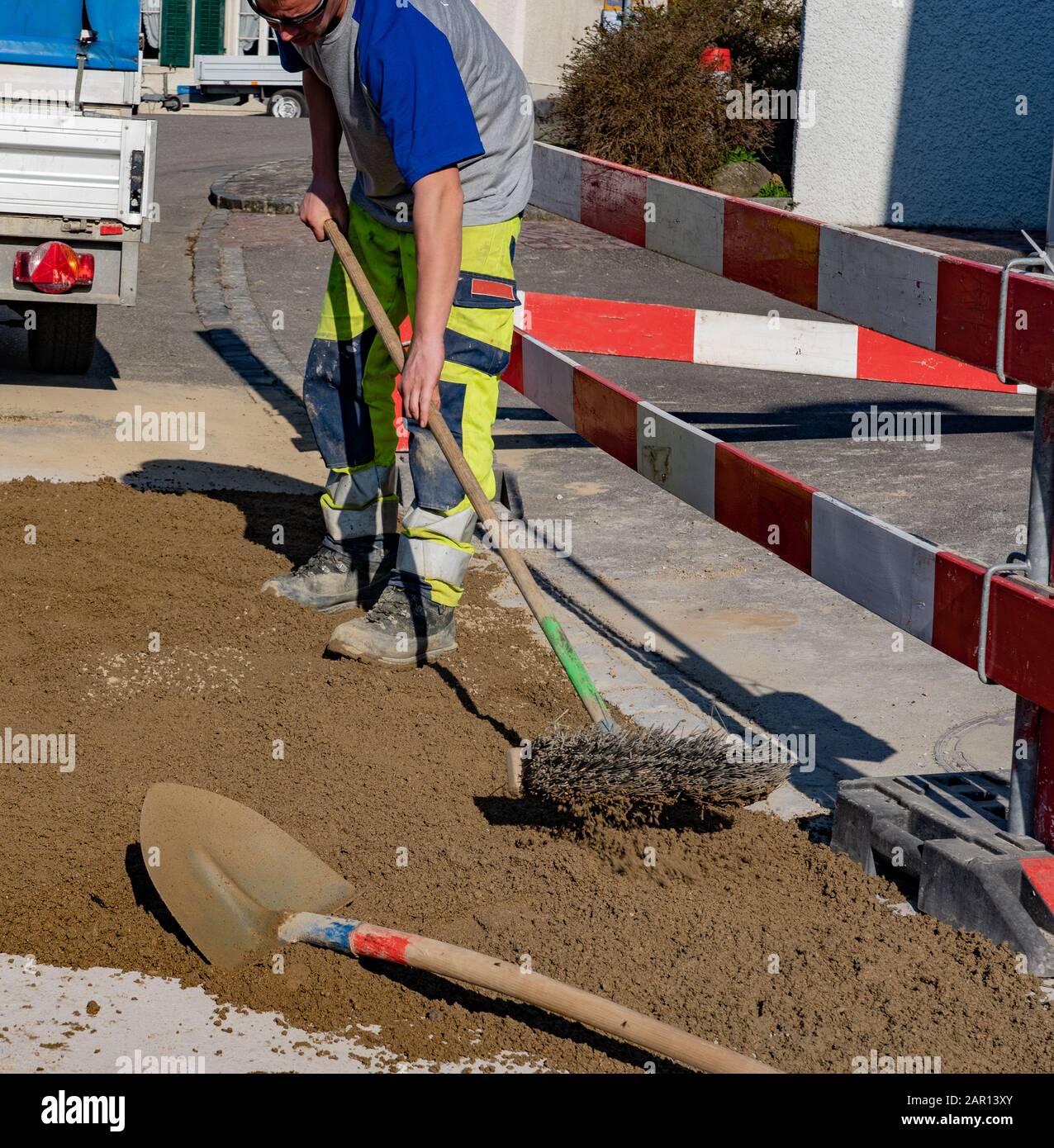 Operaio edile con occhiali da sole e spazzatrice sabbia nel cantiere di fronte a un veicolo da costruzione e rimorchio. Lavoratore edile, Foto Stock