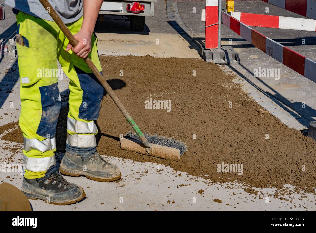 Lavoratore con pantaloni di avvertimento e scarpe da lavoro che tiene una scopa in mano e in piedi vicino alla zona di sabbia sulla strada. Cucchiaio da sabbia; pala; barri Foto Stock