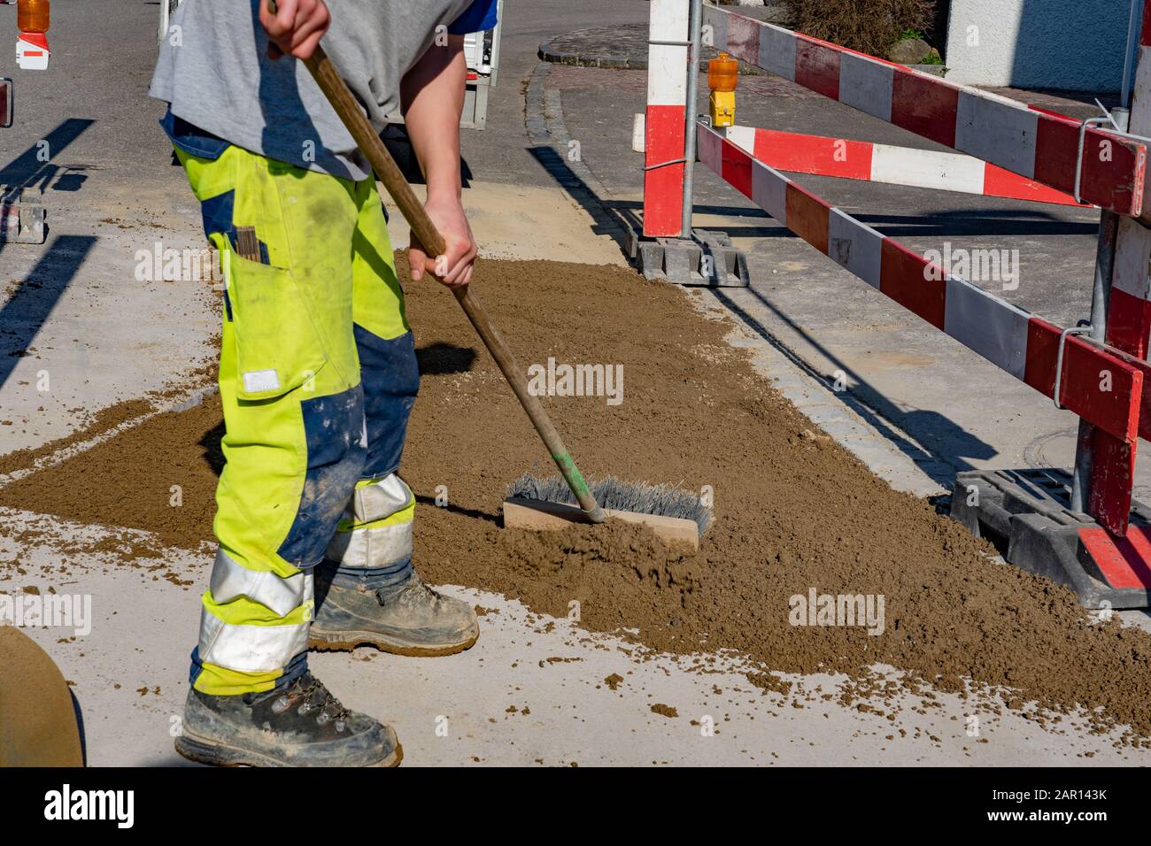 Lavoratore con pantaloni di avvertimento e scarpe da lavoro è strofinare con una scopa strada in mano sulla strada. Cucchiaio da sabbia; pala; barriera; barr. Rosso-bianco Foto Stock