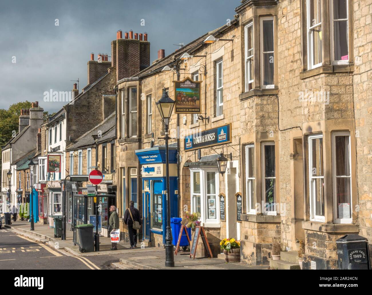 Negozi d'epoca, pub ed edifici con persone, su un lato di Galgate, nella storica città di mercato di Barnard Castle, Teesdale, County Durham, Inghilterra. Foto Stock