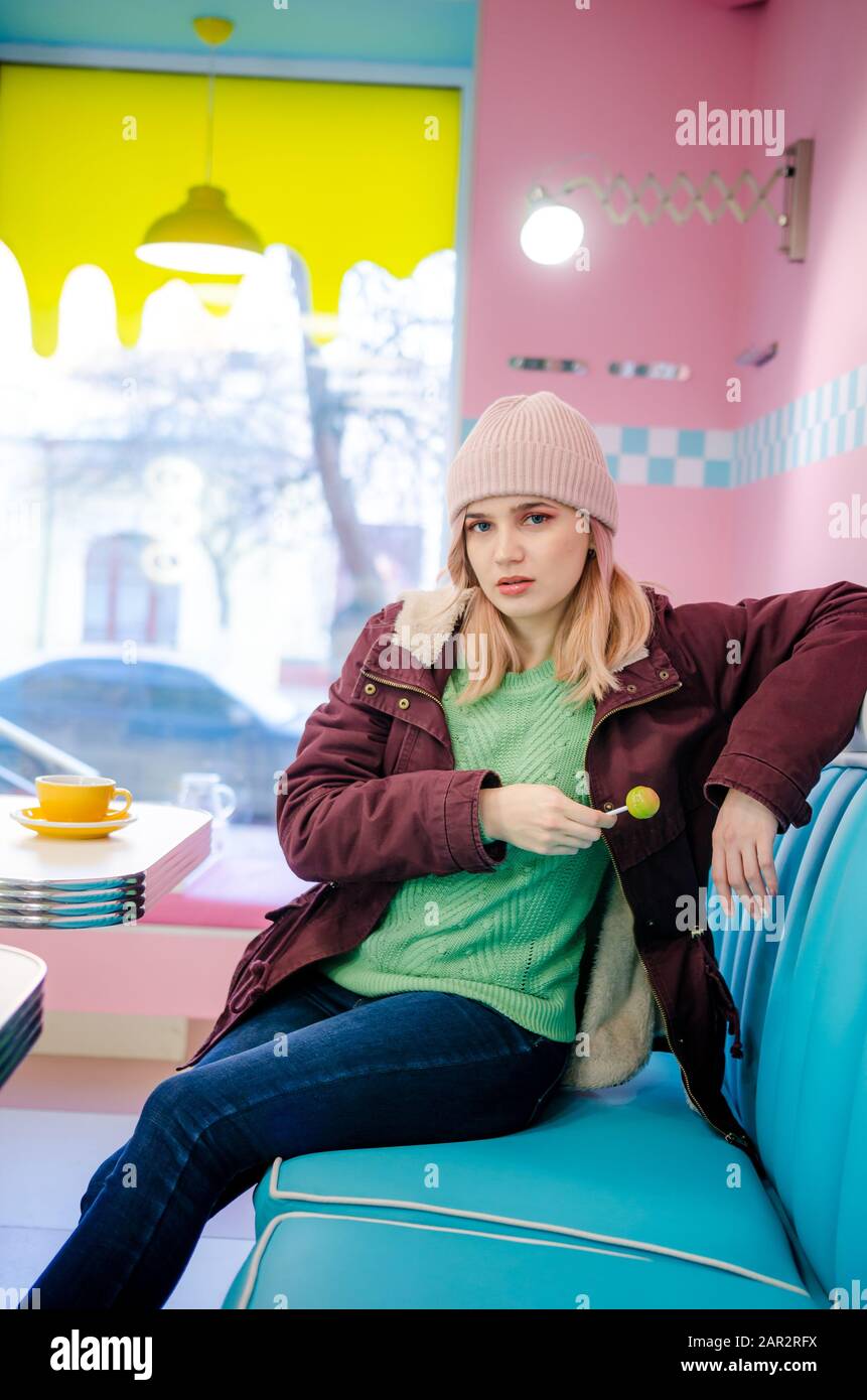 Ritratto di una ragazza bionda hippster divertente in caffè. Ragazza vestita con giacca viola, jeans e maglione verde Foto Stock