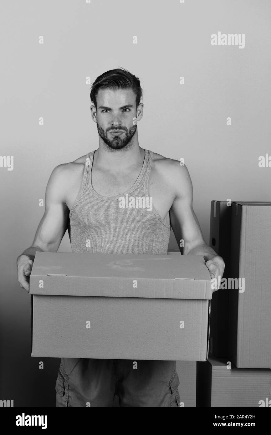 Macho con barba e viso serio. Uomo che si trova tra scatole di cartone e che ne tiene una. Ragazzo con cartone isolato su sfondo blu. Distribuzione e spostamento nel concetto. Foto Stock