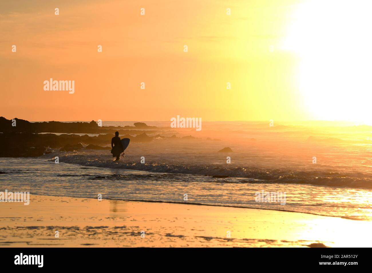 La luce solare dell'alba ardente saluta il surfista mentre entra nel surf Foto Stock