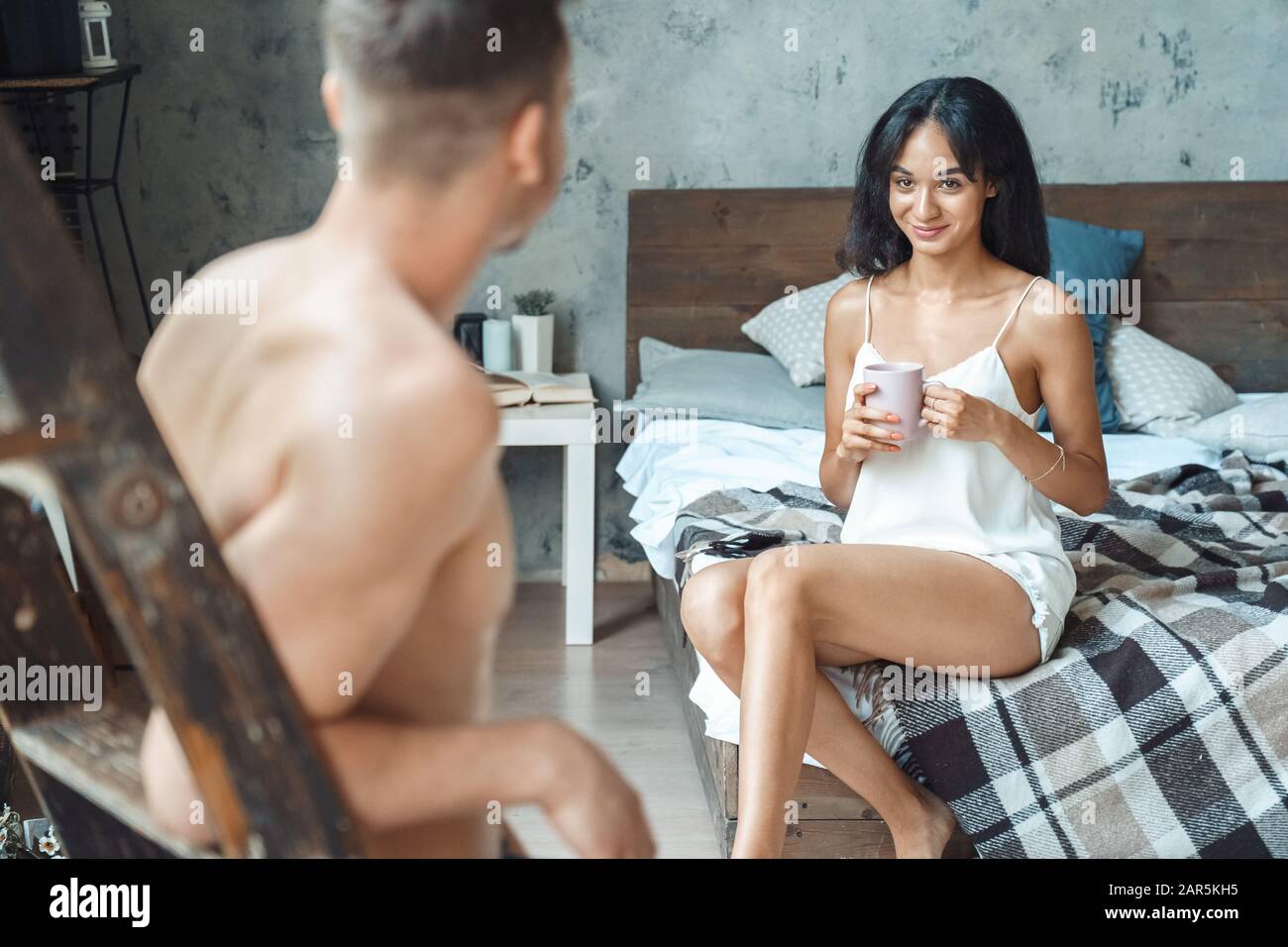 Coppia Mista Race. Giovane uomo in piedi vista sul retro vicino a guardare la donna seduto sul letto bere caffè caldo sorridente giocoso a casa Foto Stock