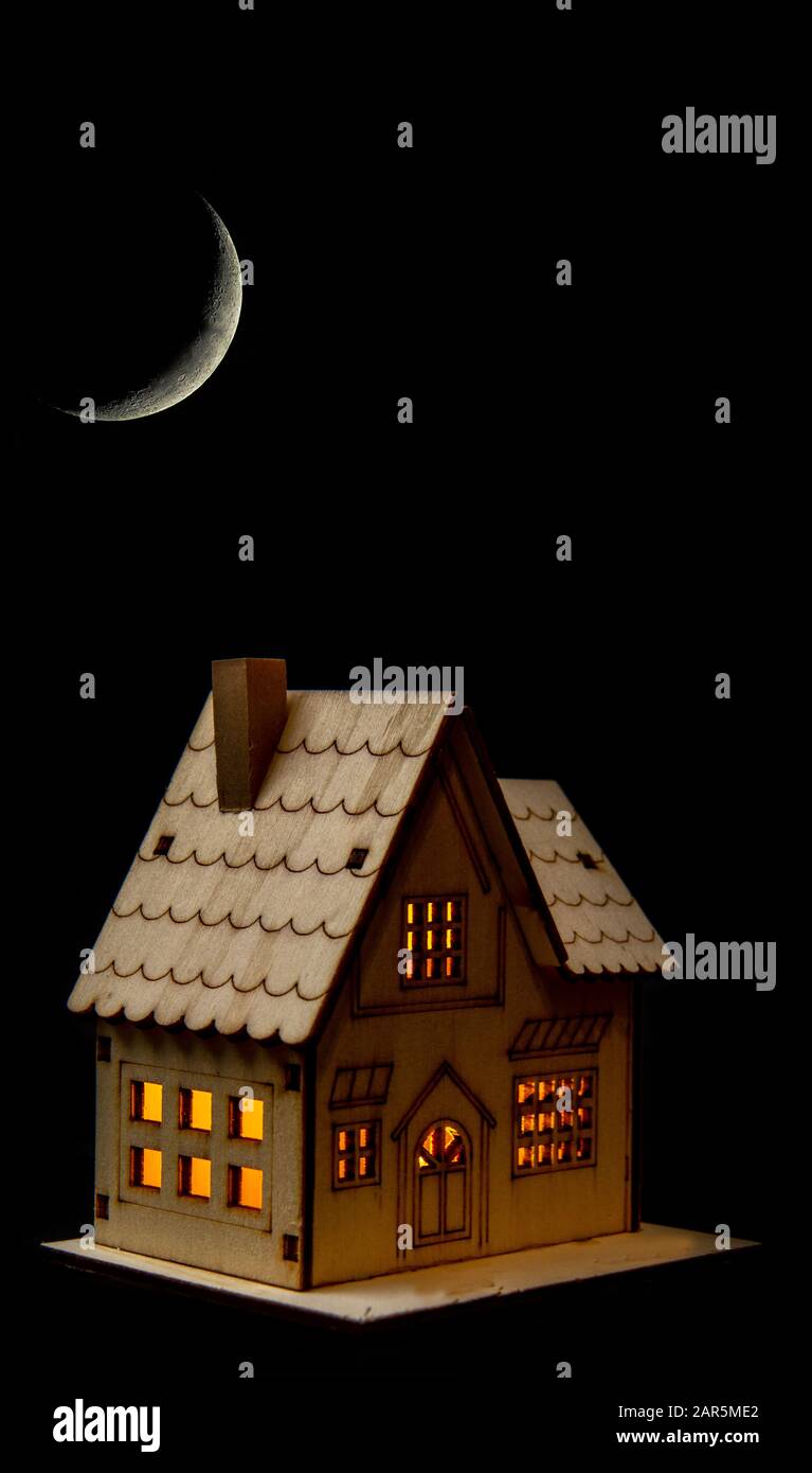 Casa giocattolo con luci accese di notte sotto una luna crescente. Lasciando le luci accese, spreco di elettricità, energia, non eco-friendly. Foto Stock