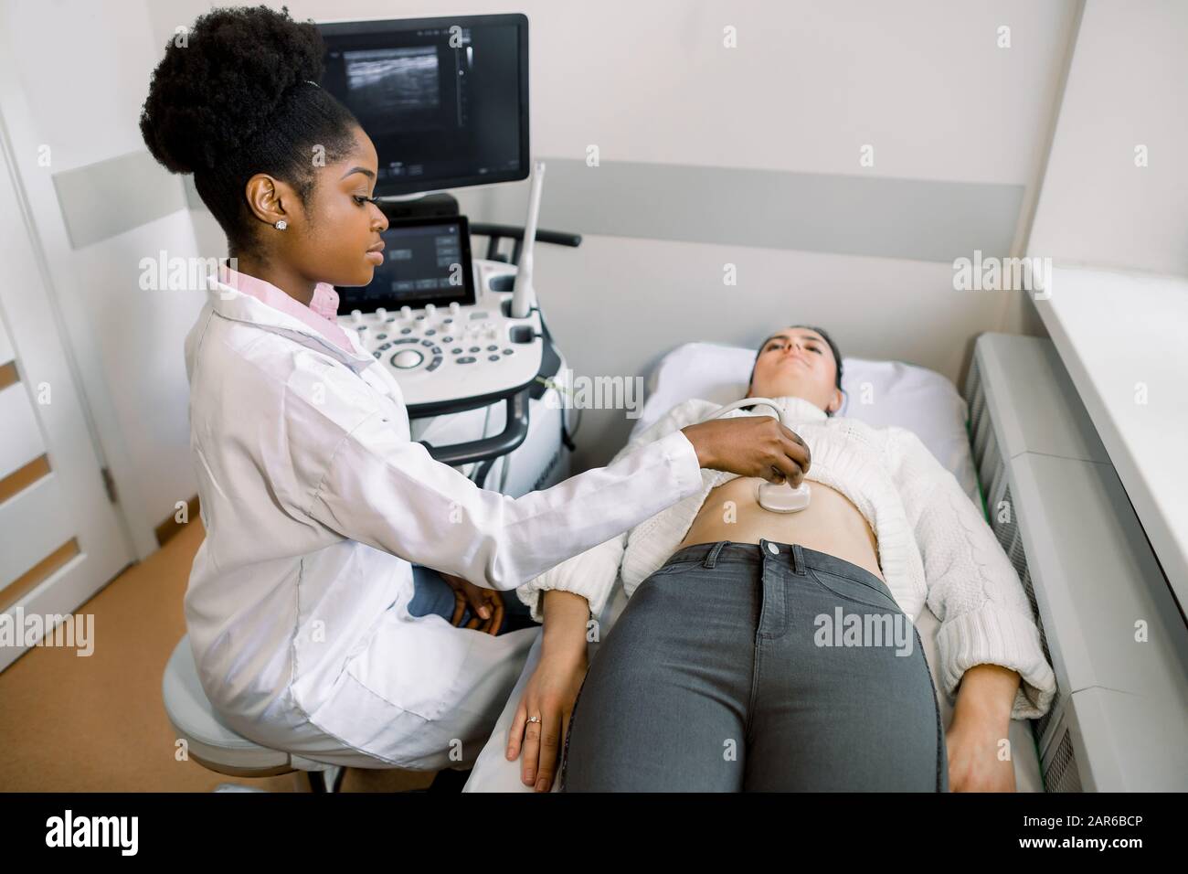 Giovane medico africano che muove il trasduttore a ultrasuoni sul ventre della donna in ospedale. Medico femminile che dà al paziente un suono ultra dell'addome. Incinta Foto Stock