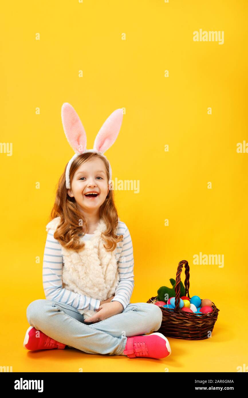 Gioiosa bambina seduta sul pavimento con un cesto di uova di Pasqua. Felice cute bella bambino in lunghezza piena nelle orecchie del coniglietto sul giallo Foto Stock