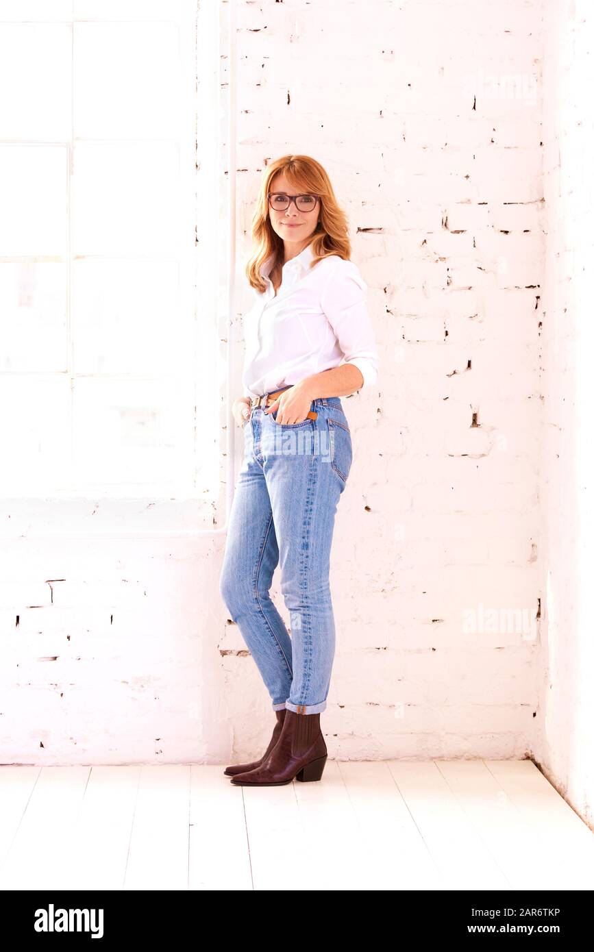 Scatto a tutta lunghezza dell'attraente donna di mezza età che indossa una camicia bianca e jeans blu mentre si rilassa al muro di mattoni bianchi. Foto Stock