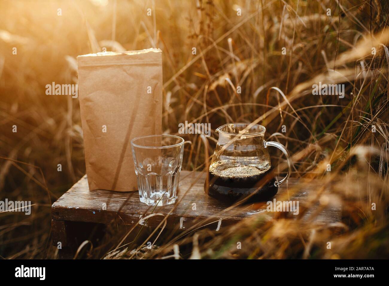 bollitore in vetro con caffè, tazza e pacchetto marrone con caffè sullo sfondo delle erbe rurali della campagna al tramonto. Caffè alternativo all'aperto. Atmo Foto Stock