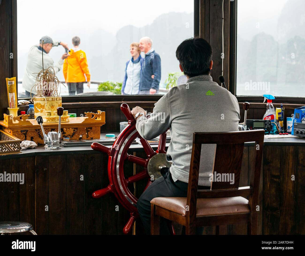 Uomo al volante del capitano in nave da crociera con i turisti che scattano foto sul ponte, Halong Bay, Vietnam, Asia Foto Stock