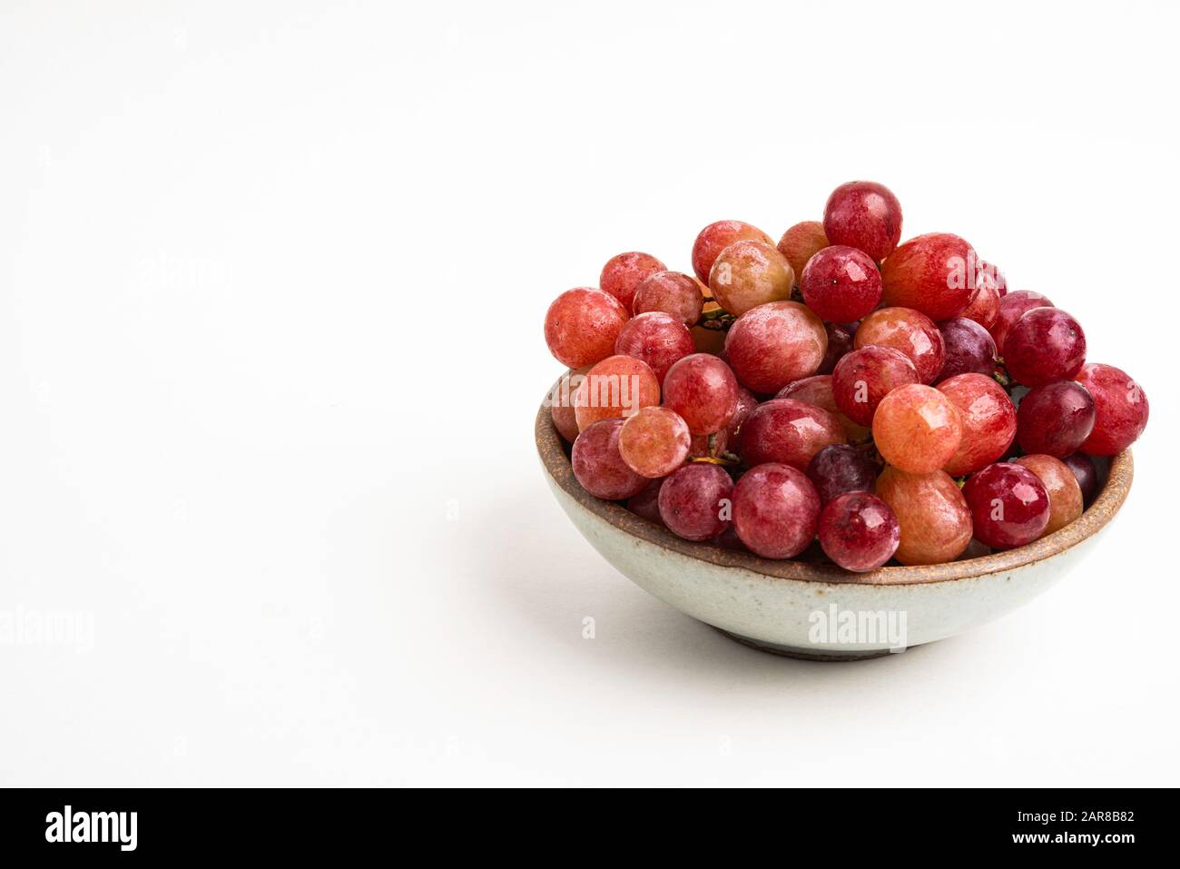 Un grappolo di uve rosse fresche raccolte su un piccolo recipiente in ceramica adagiato su uno sfondo bianco. Foto Stock
