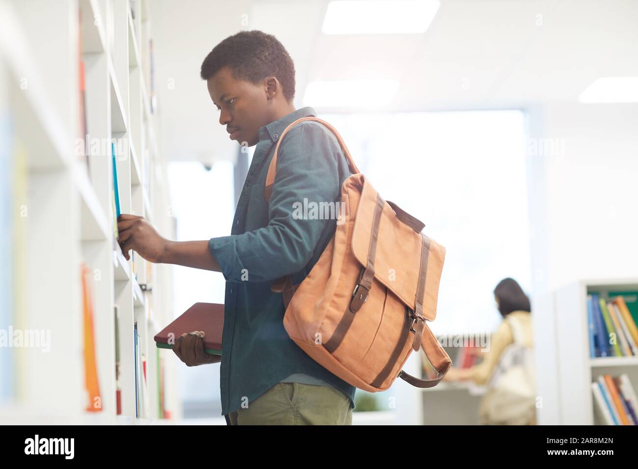 Ritratto di vista laterale dello studente africano che indossa zaino scegliendo libri sugli scaffali della biblioteca universitaria, copia spazio Foto Stock